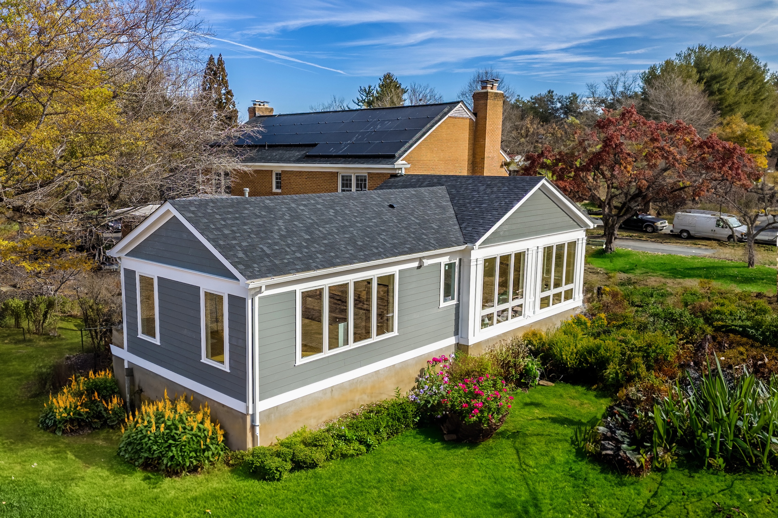 A modern sunroom with large windows and gray siding, designed by an expert ADU builder, extends from a brick house, surrounded by green grass, blooming flowers, and trees under a blue sky with wispy clouds.