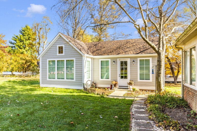 Single-story house with light gray siding, white trim, and large windows. A path bordered by plants leads to the front door. Surrounded by trees with autumn foliage, this home also features a garage conversion under a blue sky.