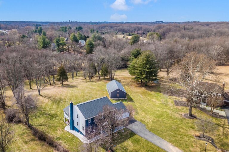 Aerial view of a house with a driveway, detached garage, and spacious lawns—plus room for an ADU or Tiny Home—scattered trees, and a forest in the background under a clear blue sky.