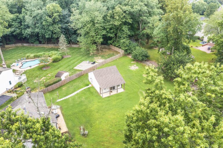 Aerial view of a large backyard with a small white shed, a fenced area containing a pool and trees, and a manicured green lawn bordered by woods and other homes.