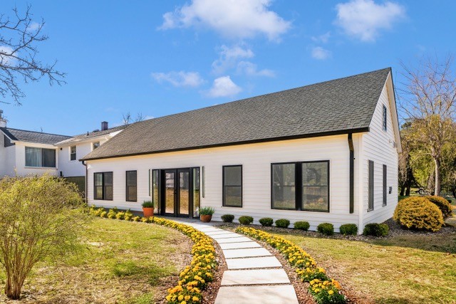 A modern white house with black trim, large windows, and a dark roof. A stone walkway lined with yellow flowers and shrubs leads to the glass front door, with a lawn and blue sky in the background.
