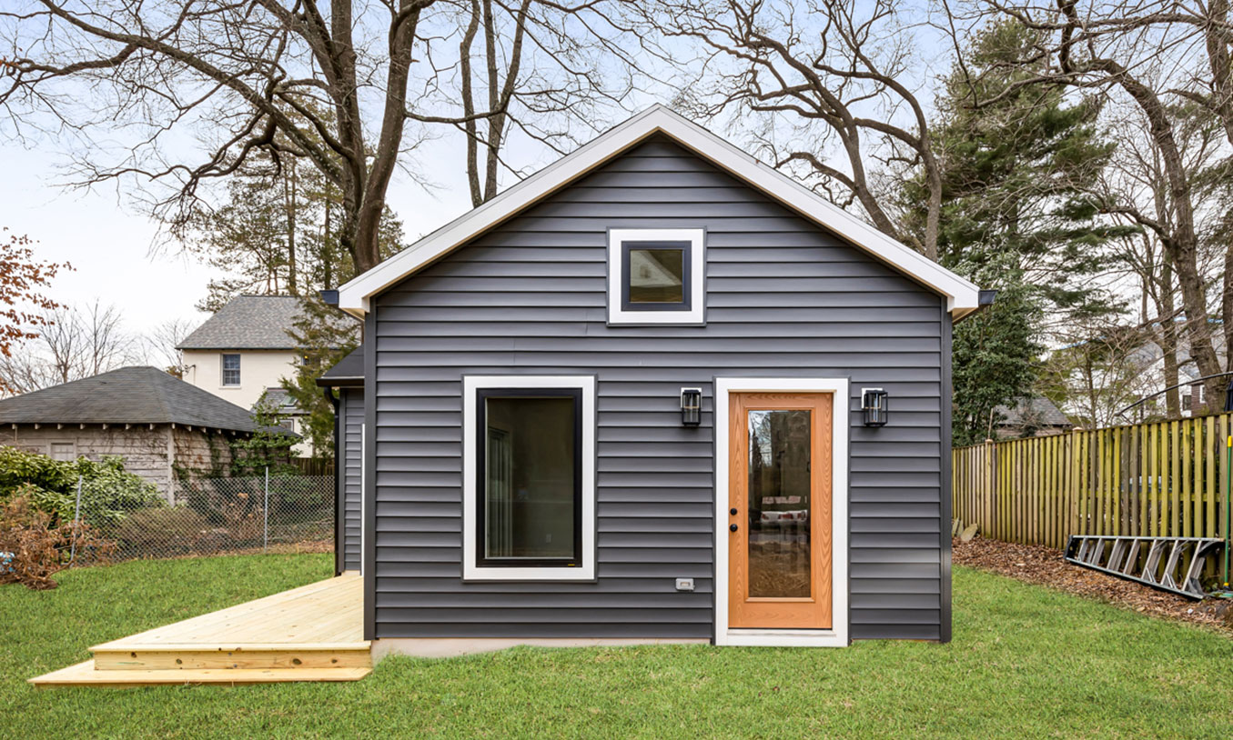 A small modern house with dark gray siding, a wooden door with a glass panel, large windows, a wooden deck on the left, and a grassy yard surrounded by trees and fencing.