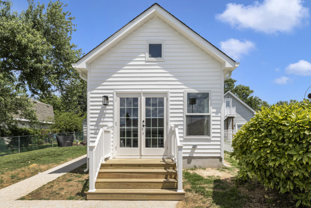 A small white house with a steep roof, glass double doors, and a single front window. Wooden steps with white railings lead to the entrance. Green lawn, trees, and blue sky with clouds surround the home.