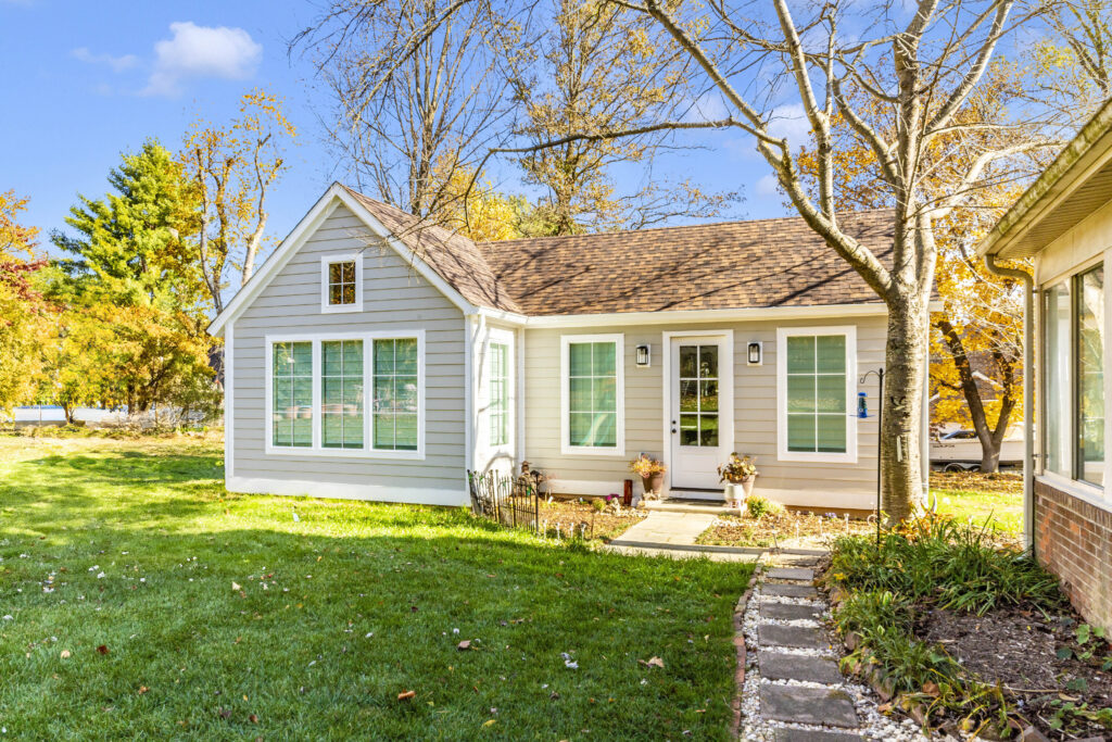 Single-story house with light gray siding, white trim, and large windows. A path bordered by plants leads to the front door. Surrounded by trees with autumn foliage, this home also features a garage conversion under a blue sky.