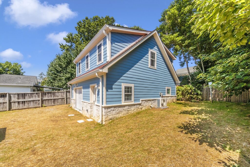 A blue two-story house with white trim and a stone foundation sits on a dry, yellowish lawn. Nearby, a Detached ADU enhances the property, both surrounded by trees and wooden fences under a clear, sunny sky.