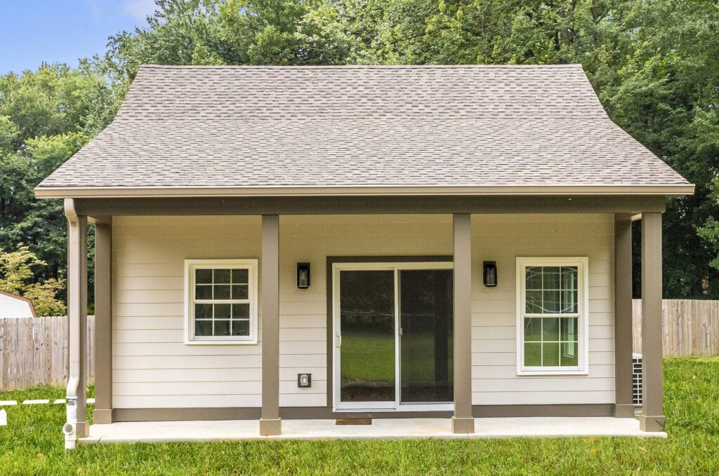 A small beige house with a sloped roof, front porch, two windows, and a glass sliding door. The house sits on a grassy lawn, surrounded by a wooden fence and trees in the background.