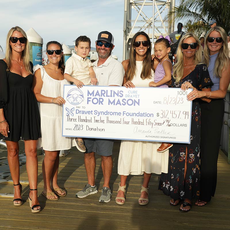 A group of seven adults and two children stand on a pier, smiling and holding a large donation check for the Dravet Syndrome Foundation from Marlins for Mason, showcasing the power of giving and charitable contributions totaling $312,457.94.
