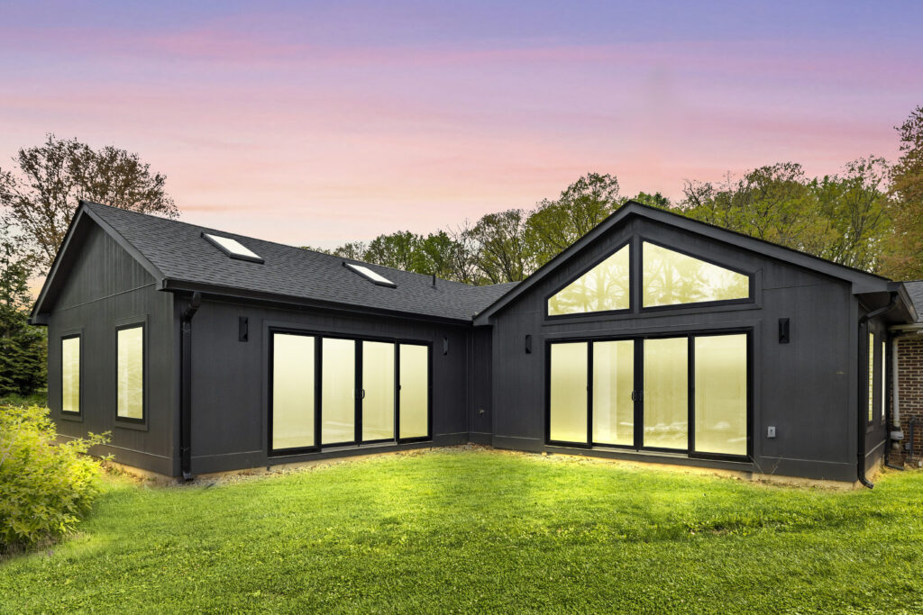 Modern black MAKARA 1100 house with large floor-to-ceiling windows glowing warmly at dusk, surrounded by green grass and trees, under a pink and purple sunset sky.
