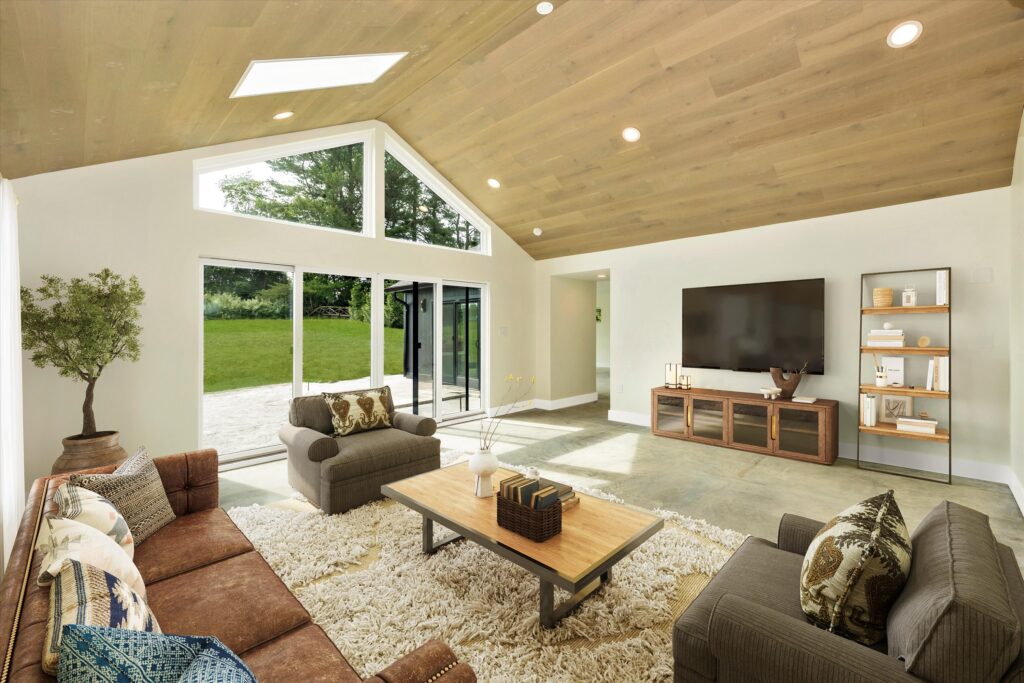 A spacious living room with a vaulted wood ceiling and large windows, featuring a MAKARA 1100 elegantly mounted above the TV. Glass doors open to a green yard, while a brown sofa set surrounds a wooden coffee table on a shag rug.