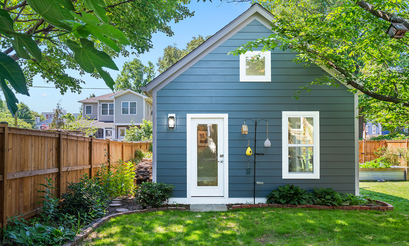 A small blue house with white trim sits in a sunny, fenced backyard surrounded by green trees and plants. There are two windows and a glass door facing the yard, with a neighboring house visible in the background.
