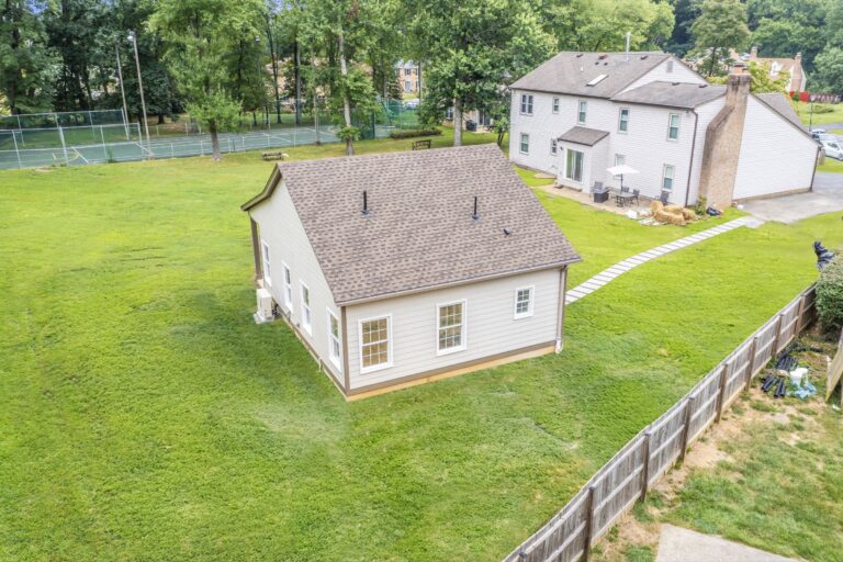 A small, white detached building with windows sits on a grassy lawn near a larger house. A tennis court and tall trees are visible in the background, and a wooden fence surrounds part of the property.